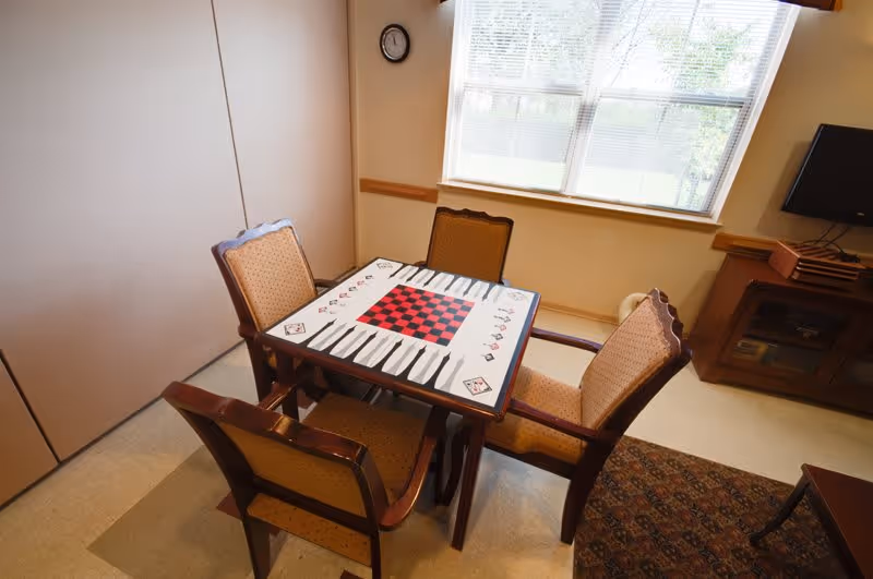 A small room with a square table designed for playing board games such as checkers and backgammon, surrounded by four cushioned wooden chairs. The room has a window with blinds partially open, a wall clock, a TV on a wooden stand, and a patterned rug on the floor.