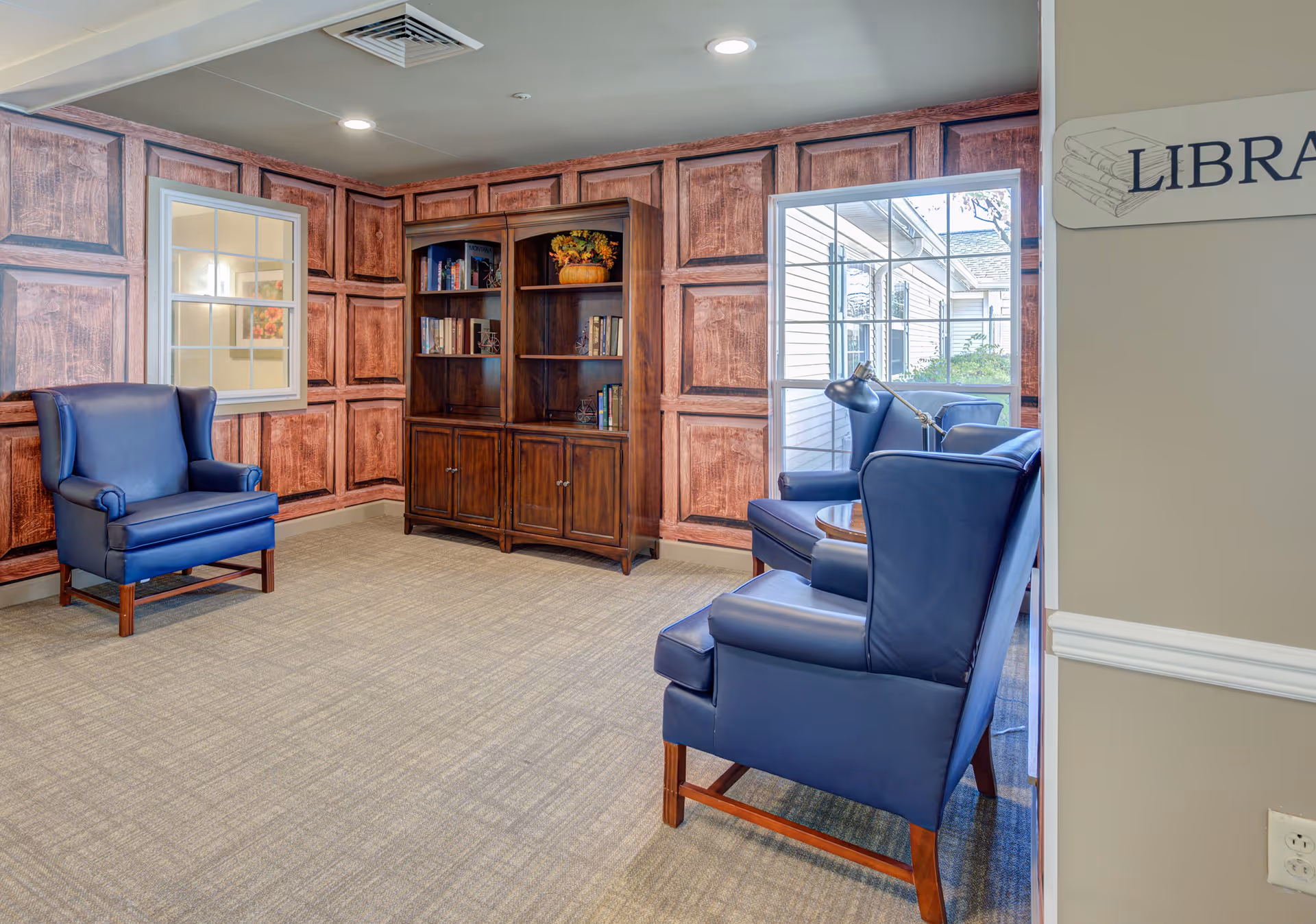 A cozy library room with three blue leather armchairs arranged around a small wooden table with a reading lamp. There is a wooden bookshelf filled with books and decorative items against a wall with a wood panel design. A window shows an outdoor view, and a sign on the wall reads 'LIBRARY'.