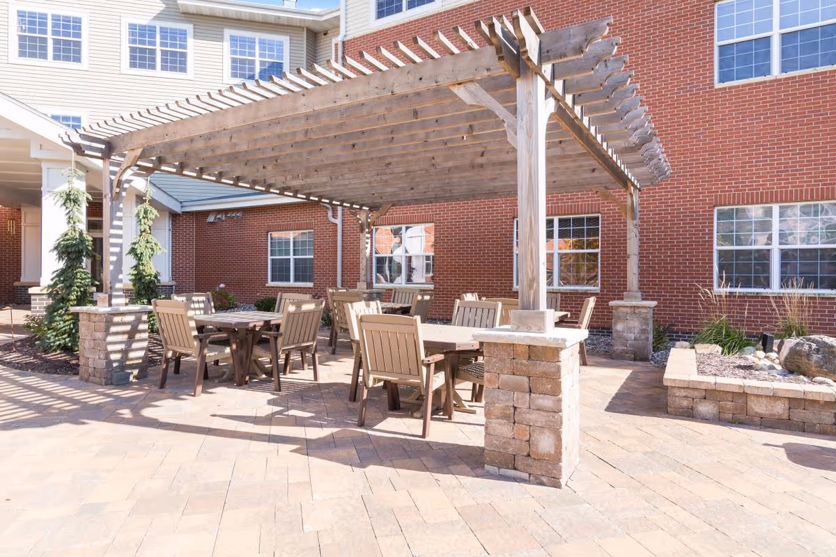 Outdoor patio courtyard with a wooden pergola, tables and chairs in front of a brick senior living building.
