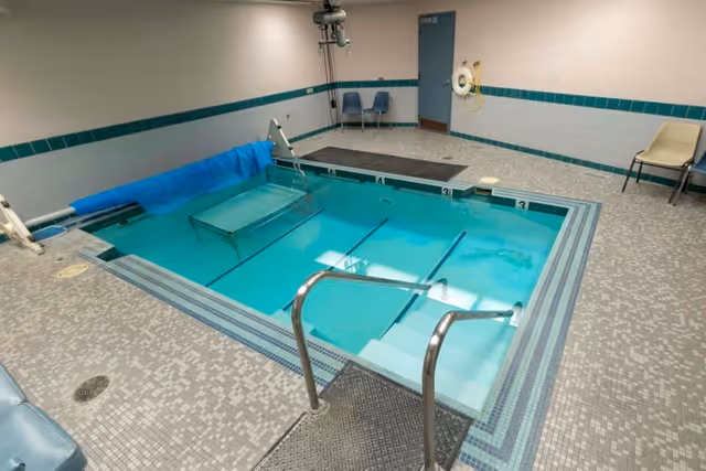 Indoor therapeutic pool with clear blue water, metal handrails, steps leading into the pool, and a pool lift. The room has tiled floors and walls with a teal border, two chairs against the wall, and a life preserver mounted on the wall.