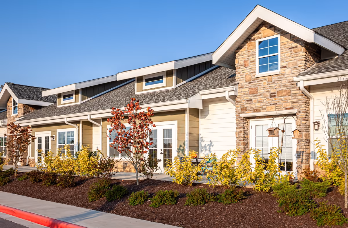 Exterior view of a senior living facility building with beige siding, stone accents, multiple windows, and a landscaped garden with small trees and shrubs under a clear blue sky.