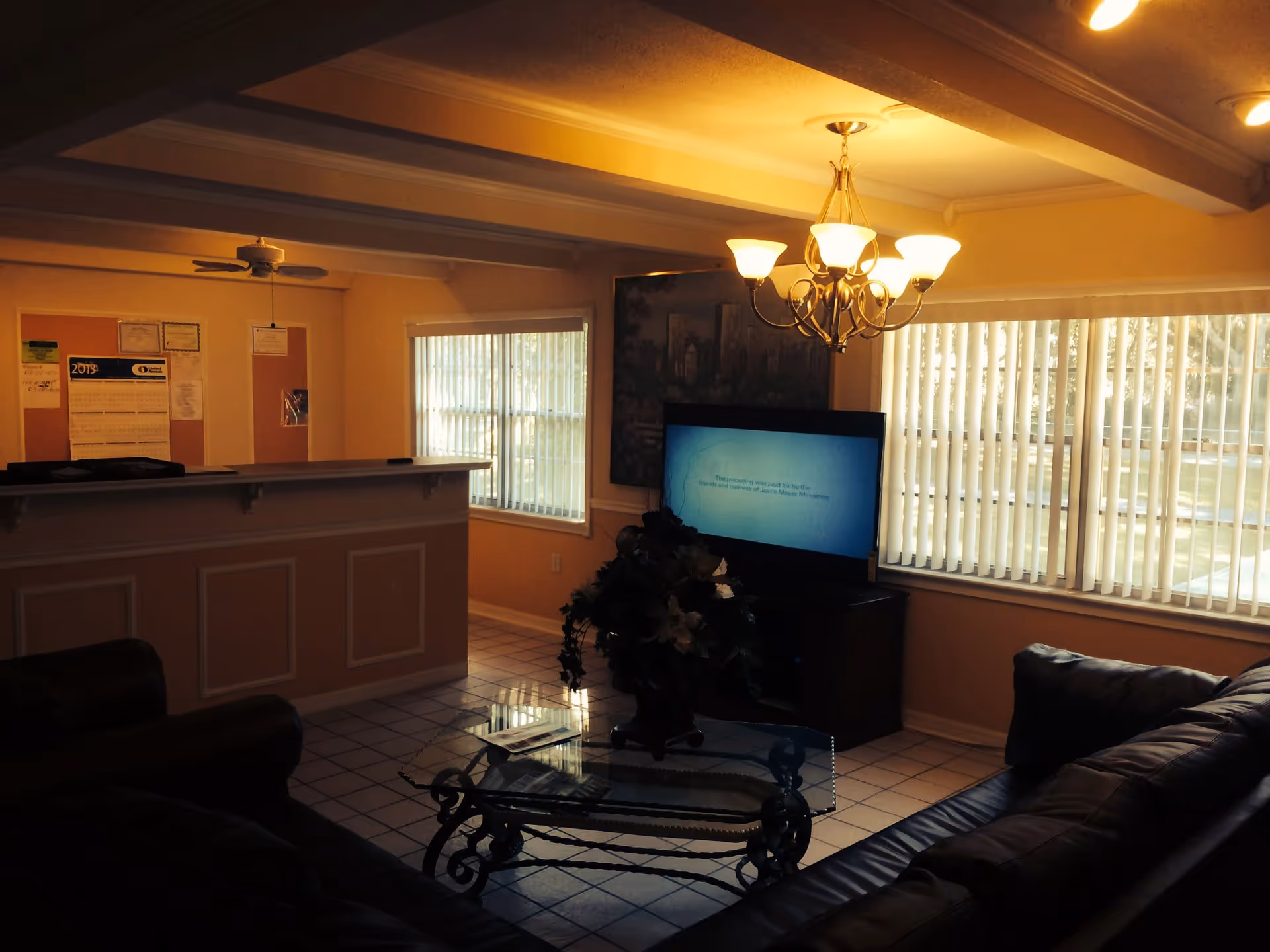 Interior view of a senior living facility common area with a leather couch, glass coffee table with a floral arrangement, a flat-screen TV, and a reception desk with bulletin boards on the wall behind it. The room has tiled floors, large windows with vertical blinds, and a chandelier hanging from the ceiling.