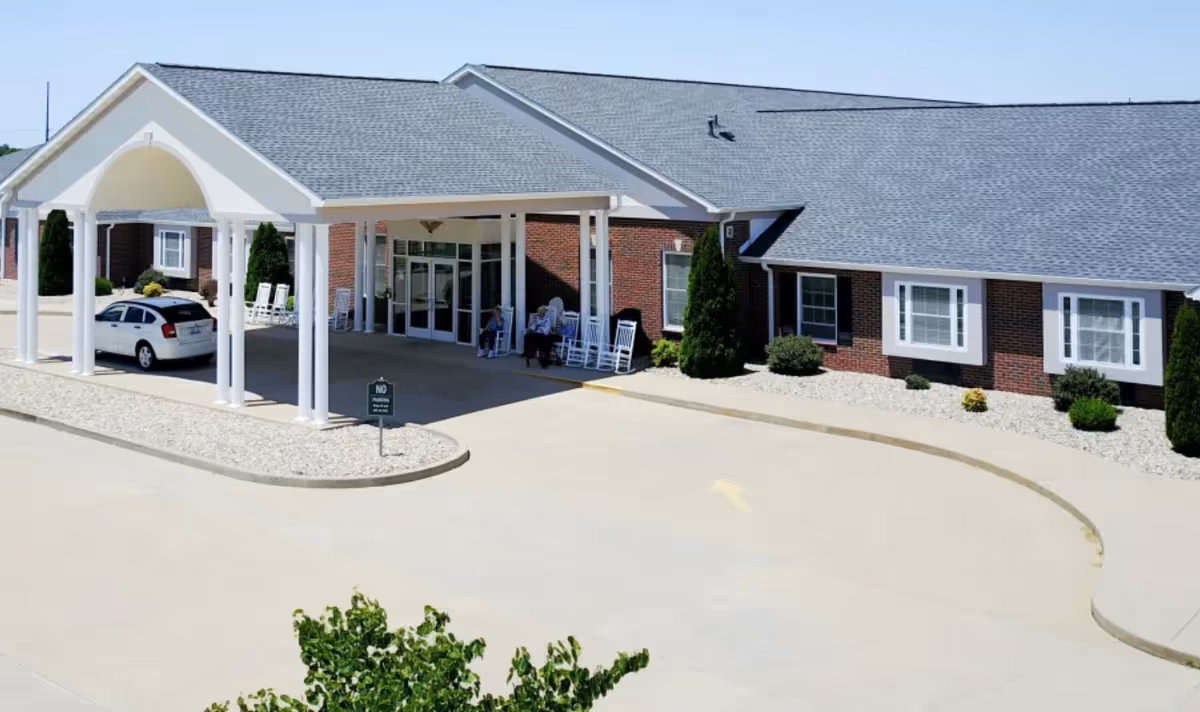 Front entrance of a single-story brick senior living facility with a covered porte-cochere, rocking chairs and a parked car.