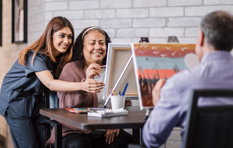 A young woman in scrubs assists an older woman with painting on a canvas at a table, while a man in a purple shirt paints on another canvas nearby in a bright room with a brick wall background.