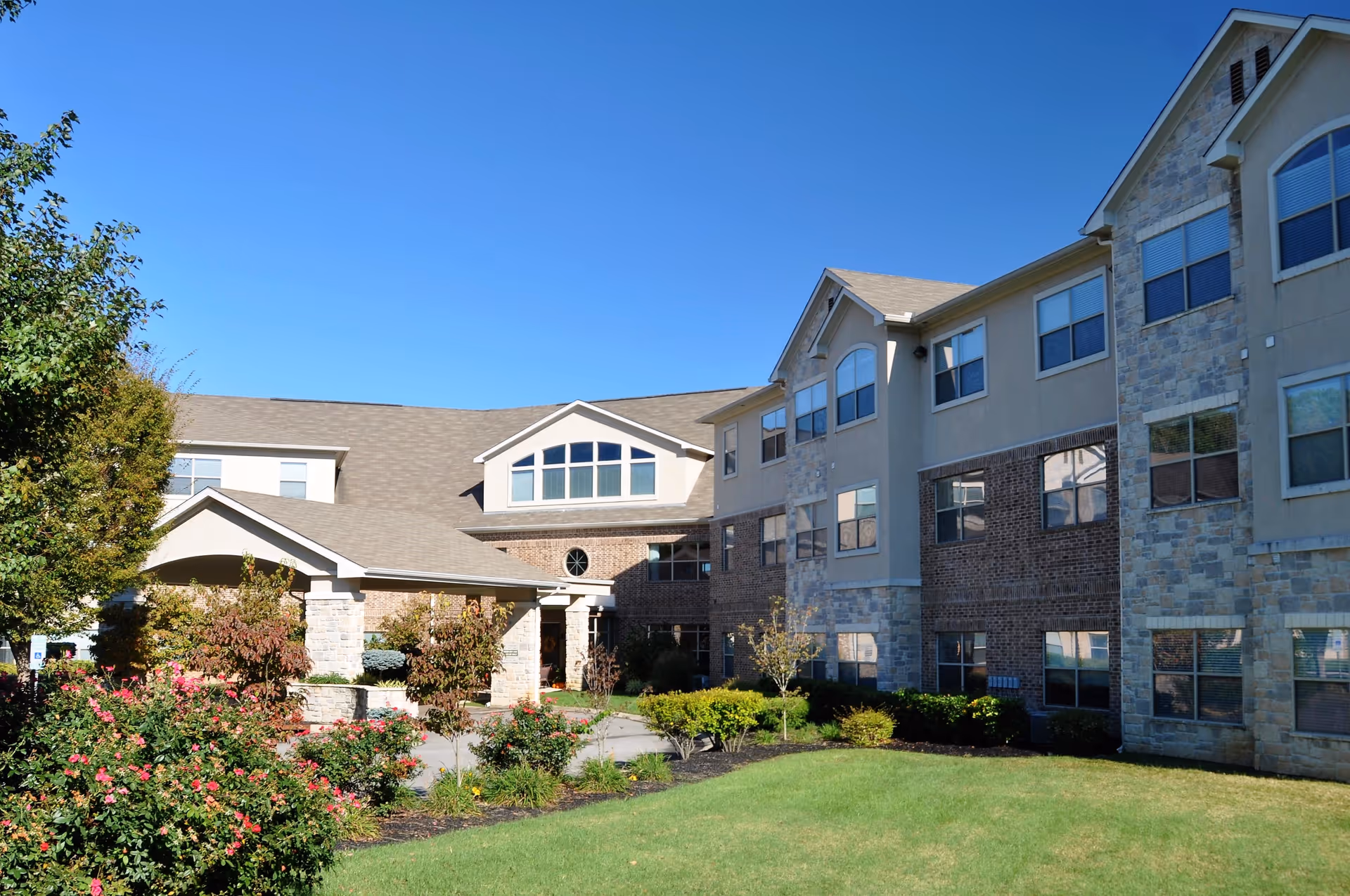 Exterior view of a senior living facility building with a well-maintained lawn, bushes, and flowering plants under a clear blue sky.