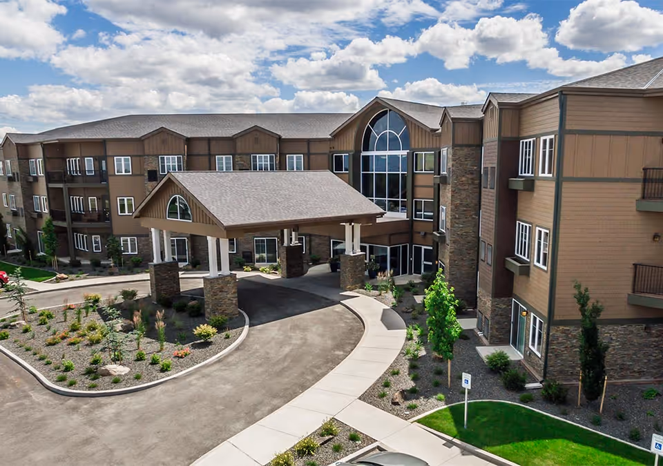 Exterior view of a large senior living facility building with multiple floors, featuring a covered entrance supported by stone pillars, landscaped gardens, and a curved driveway under a partly cloudy sky.