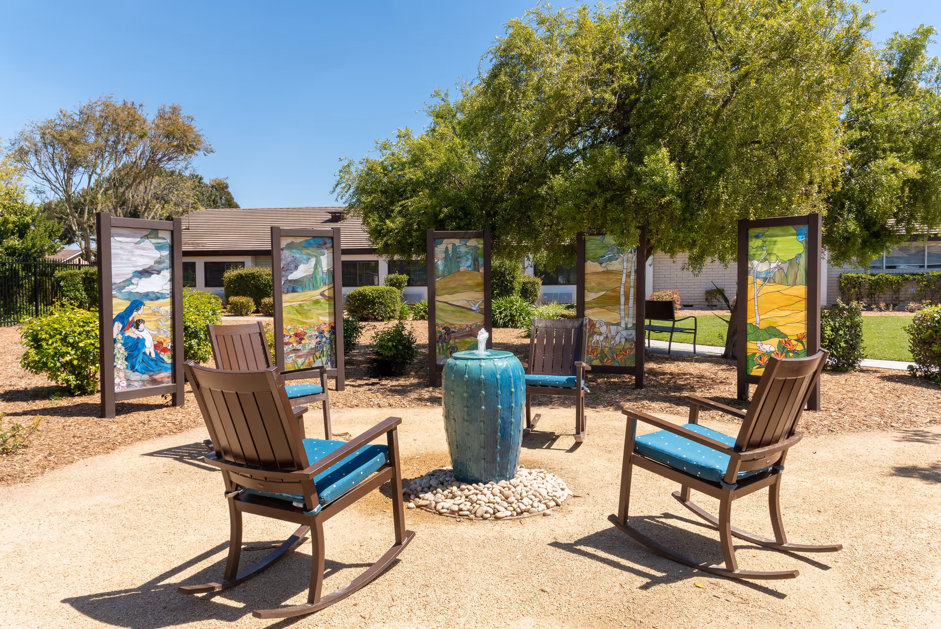 Outdoor seating area with four wooden rocking chairs with blue cushions arranged around a small blue water fountain on a gravel surface. Behind the chairs are five decorative panels with colorful landscape and nature-themed artwork. The area is surrounded by greenery, bushes, and trees, with a building visible in the background under a clear blue sky.