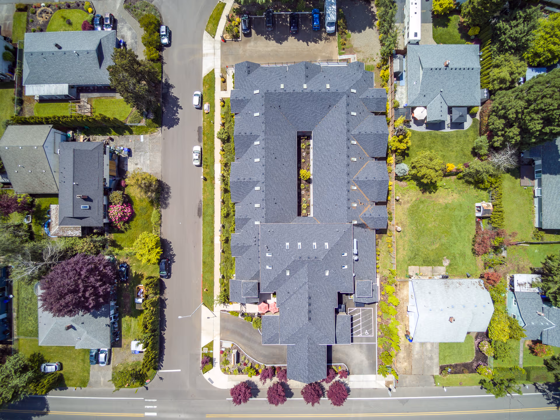 Aerial view of River Terrace Memory Care facility showing a large building with a central courtyard, surrounded by streets, parked cars, and neighboring houses with green lawns and trees.