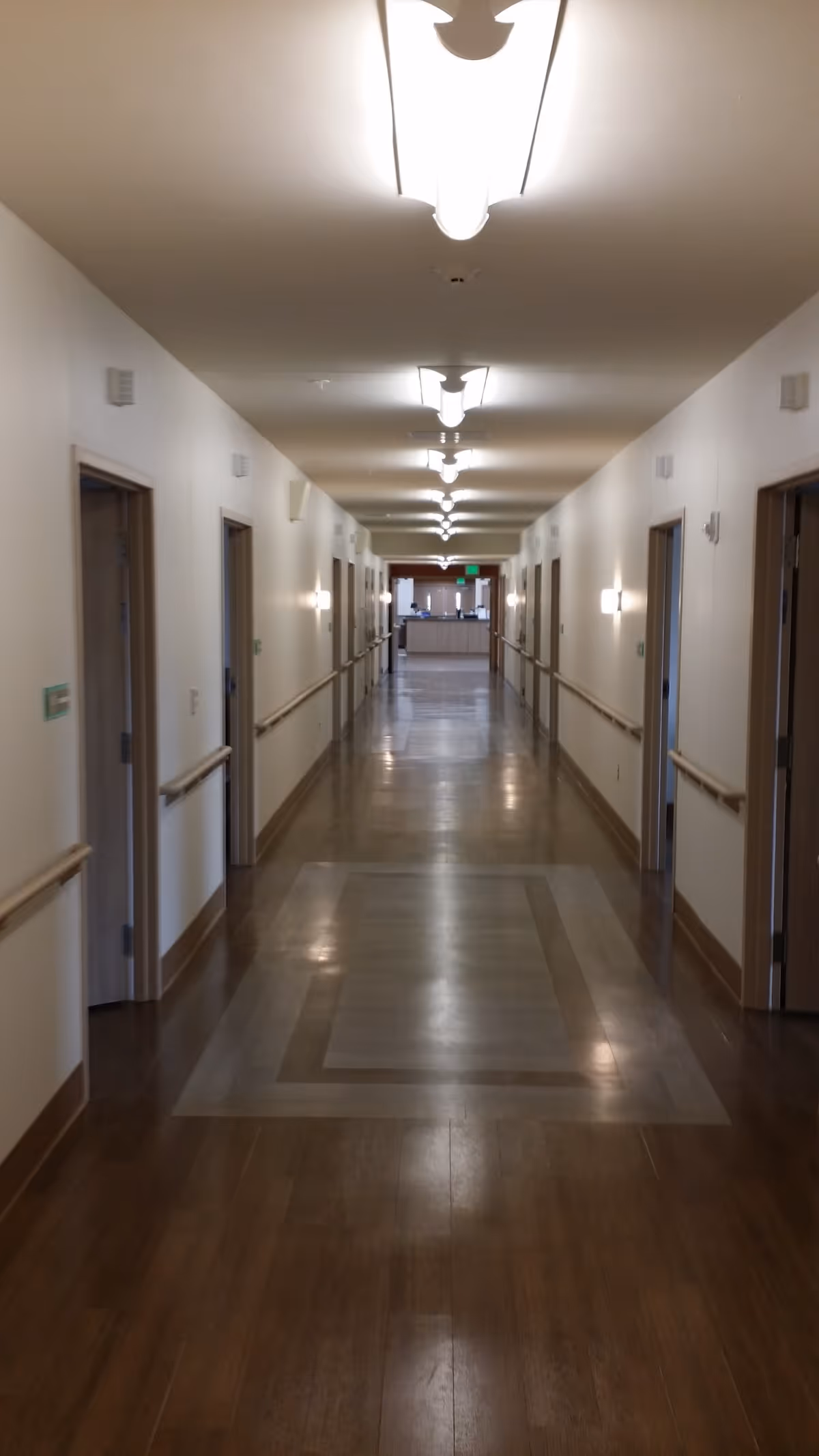 A long, well-lit hallway in a senior living or rehab facility with wooden floors and white walls. There are multiple doorways on both sides of the hallway, handrails along the walls, and ceiling lights evenly spaced. At the end of the hallway, there is a reception or nurse station area.