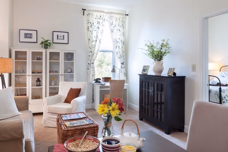 A bright and cozy living room with a beige sofa, a white armchair with a brown pillow, a wicker coffee table, and a black cabinet with a vase of green plants on top. A window with floral curtains lets in natural light. A small desk with a chair is placed near the window. In the foreground, a table holds a bowl of cereal, a cup of coffee, a glass of orange juice, and a teapot. A doorway leads to a bedroom with a bed visible.