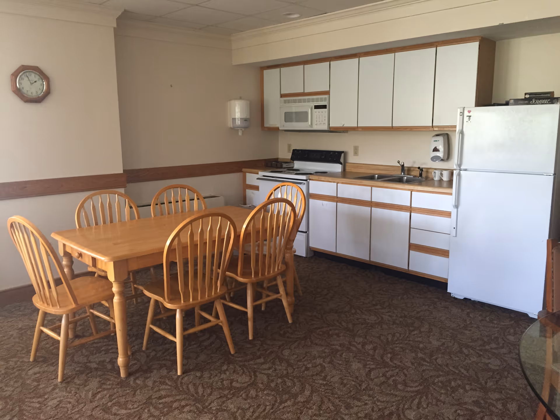A kitchen and dining area with a wooden table and six matching wooden chairs. The kitchen has white cabinets with wooden trim, a white refrigerator, a white stove with an oven, a microwave, and a double sink. There is a wall clock and a paper towel dispenser mounted on the wall. The floor is carpeted with a patterned design.