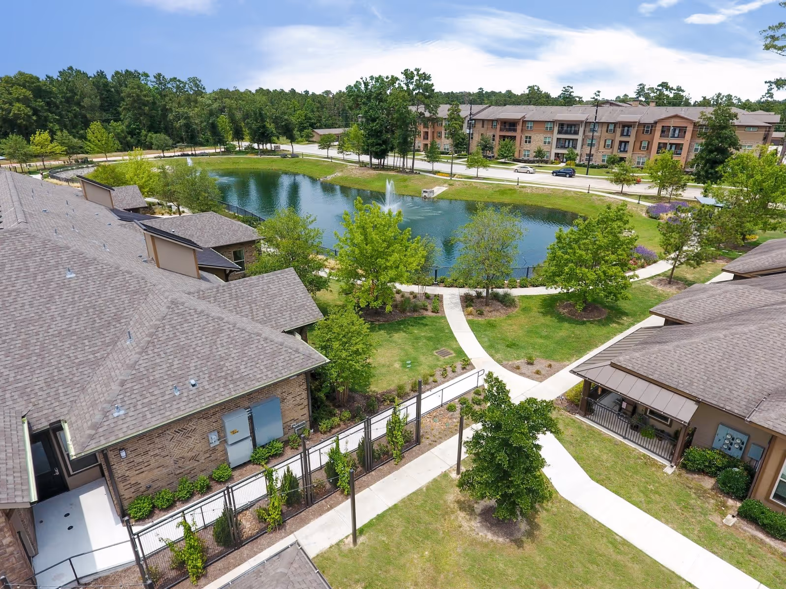 Aerial view of Watercress Assisted Living facility showing multiple buildings with brown roofs surrounding a landscaped area with a pond featuring a fountain. There are paved walkways, green lawns, trees, and shrubs around the pond and buildings. In the background, there is a road and additional residential buildings.