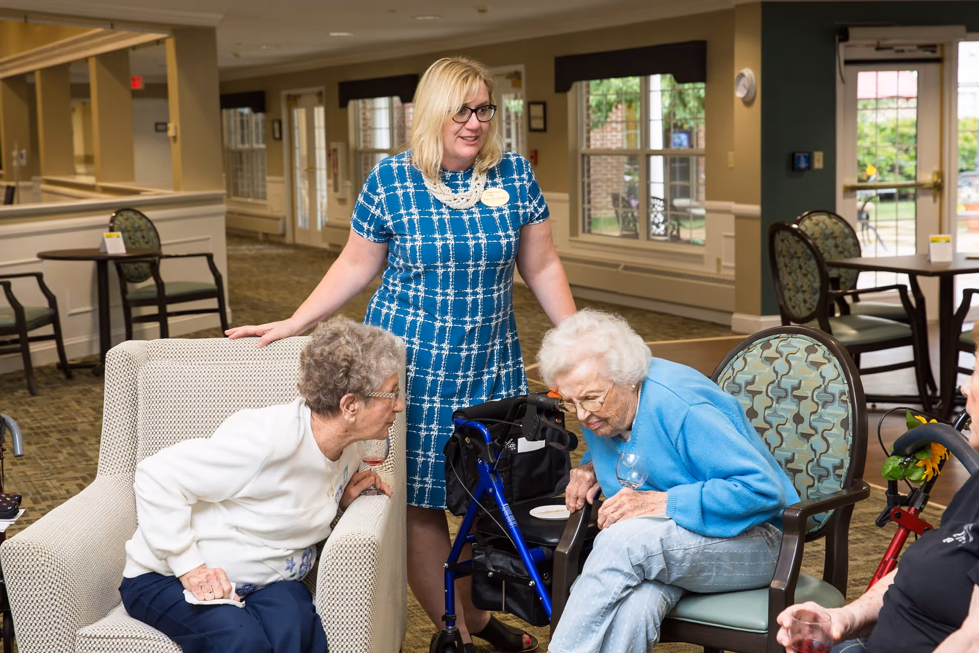 A woman in a blue patterned dress stands and smiles while interacting with two elderly women seated in armchairs in a well-lit common area with tables and chairs. One elderly woman is wearing a white sweater and glasses, and the other is wearing a blue sweater and holding a glass. A blue walker is positioned between them.
