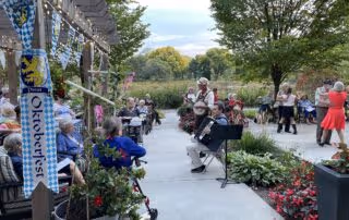 Outdoor patio area at Parkshore Senior Campus with several elderly people sitting and socializing. Some are seated under a pergola decorated with Oktoberfest banners, while others are gathered around tables and walking along a paved path surrounded by greenery and flowers.