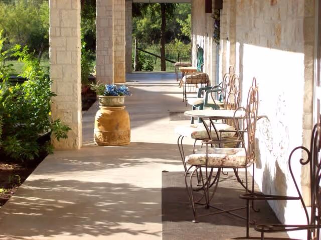 Covered stone porch with metal chairs and small tables lined along a building, potted plants, and sunlight.
