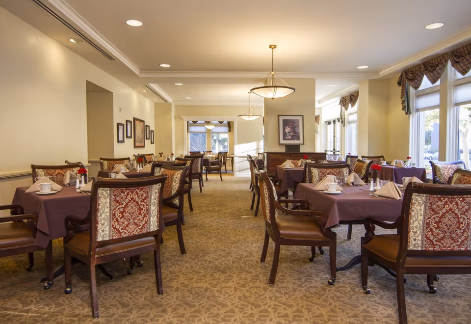 A well-lit dining room with multiple tables covered in purple tablecloths, each set with white cups, napkins, and small flower vases. The chairs have ornate upholstery with a red and beige pattern. Large windows on the right side allow natural light to fill the room, and framed artwork decorates the walls.