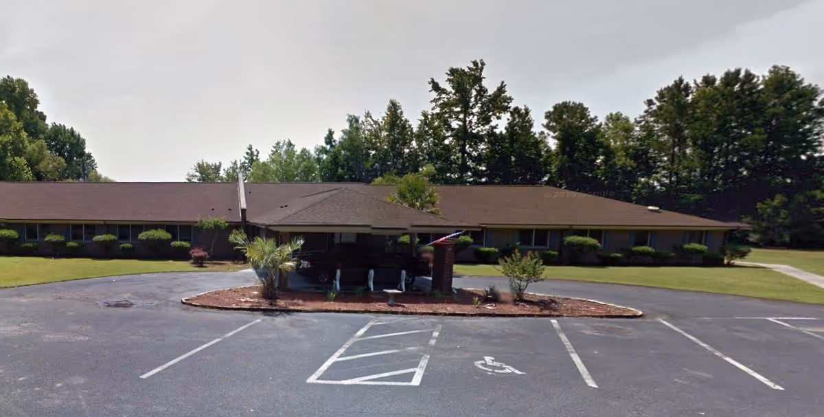 Single-story building with a brown roof and brick exterior surrounded by trees and bushes. In front of the building is a circular driveway with a covered entrance and a small landscaped area with plants. There are several empty parking spaces, including a handicapped parking spot, in the foreground.