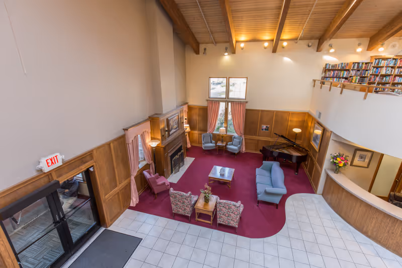 A spacious senior living facility common area with a high wooden ceiling and large windows. The room features a red carpeted seating area with various chairs and sofas arranged around a coffee table, a fireplace, a grand piano, and bookshelves on the upper level. There is a tiled floor near the entrance with glass doors and an exit sign above them.