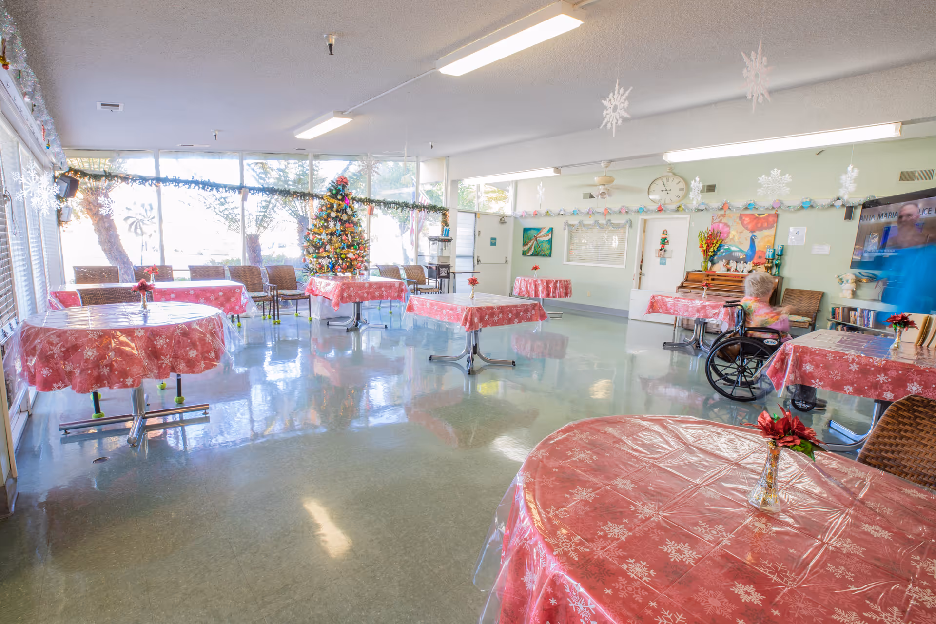 A bright and spacious common room decorated for Christmas with several round tables covered in red tablecloths with white snowflake patterns. A decorated Christmas tree stands near the large windows letting in natural light. There are chairs around the tables, a piano against the far wall, and a person in a wheelchair facing a television mounted on the wall. Snowflake decorations hang from the ceiling.