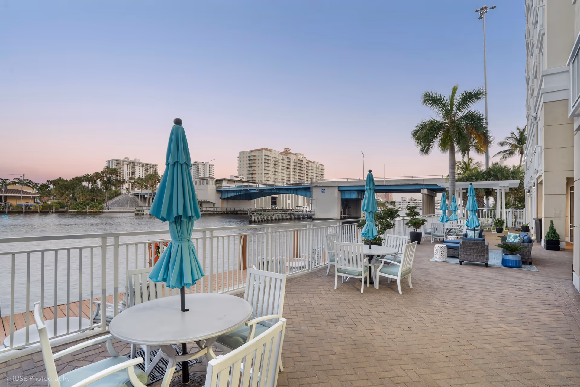 Outdoor patio area at The Meridian at Waterways featuring round tables with white chairs and closed turquoise umbrellas. The patio overlooks a body of water with a bridge and buildings in the background. Palm trees and outdoor seating with cushions are also visible.