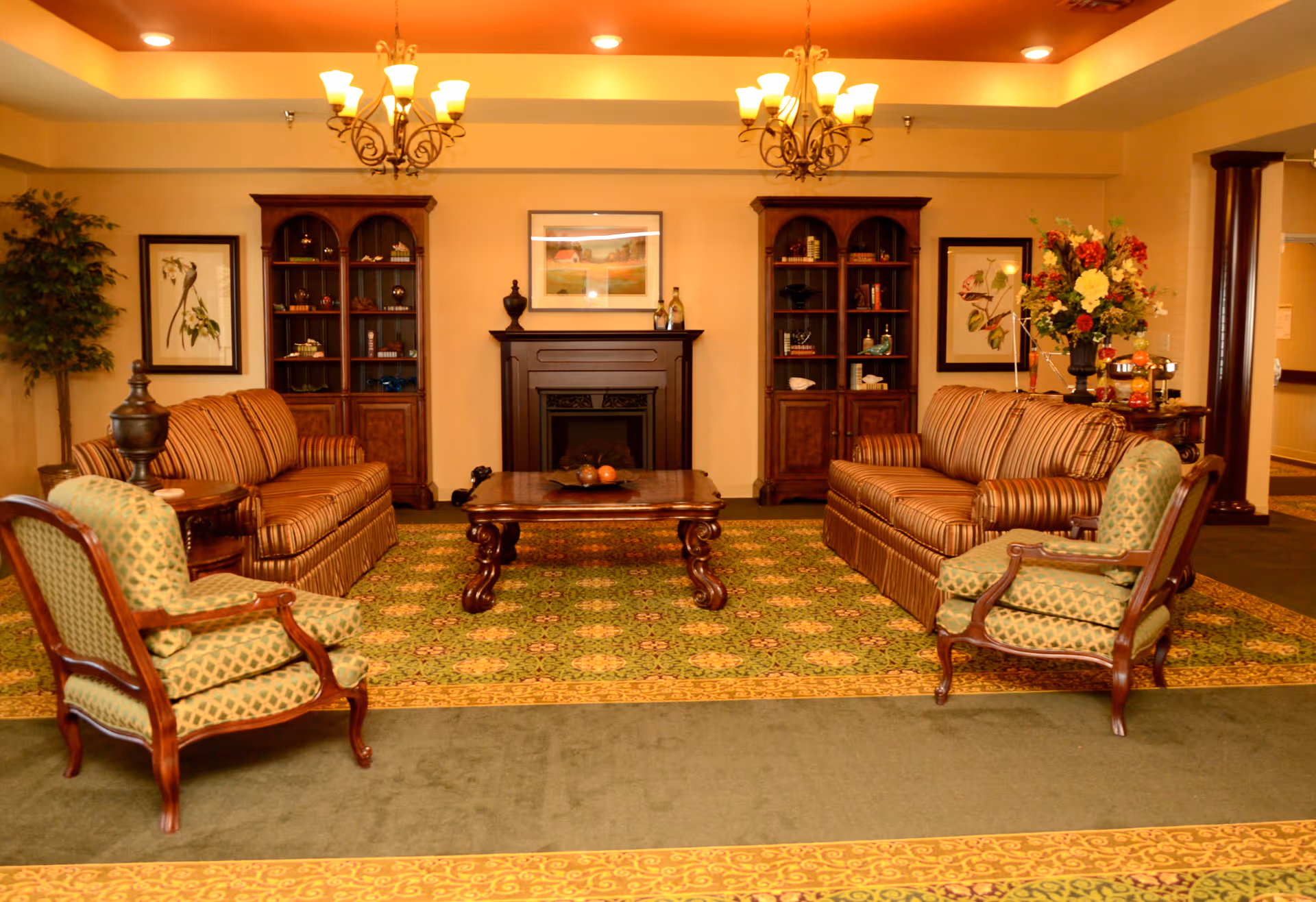 A cozy living room area with two striped sofas facing each other, two green patterned armchairs, a wooden coffee table in the center, and a fireplace with a framed picture above it. There are two wooden bookshelves on either side of the fireplace, decorative plants, and warm lighting from chandeliers on the ceiling.