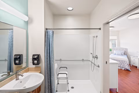 A clean, accessible bathroom featuring a white sink with a silver faucet, a large mirror, and a walk-in shower with grab bars and a white shower chair. The shower has a blue curtain and a handheld showerhead. Through an open doorway, a bedroom with a bed, nightstand, and window is visible.