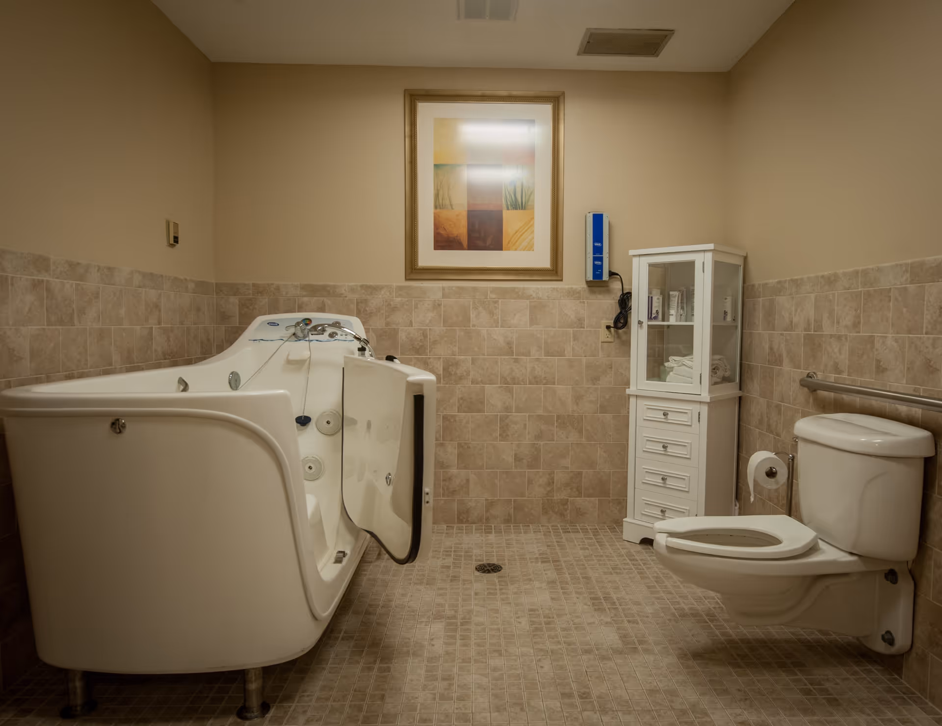 A bathroom with beige tiled walls and floor, featuring a walk-in bathtub with an open door on the left, a white toilet with a grab bar on the right, and a white cabinet with glass doors containing towels and toiletries. A framed abstract artwork hangs on the wall above the bathtub.
