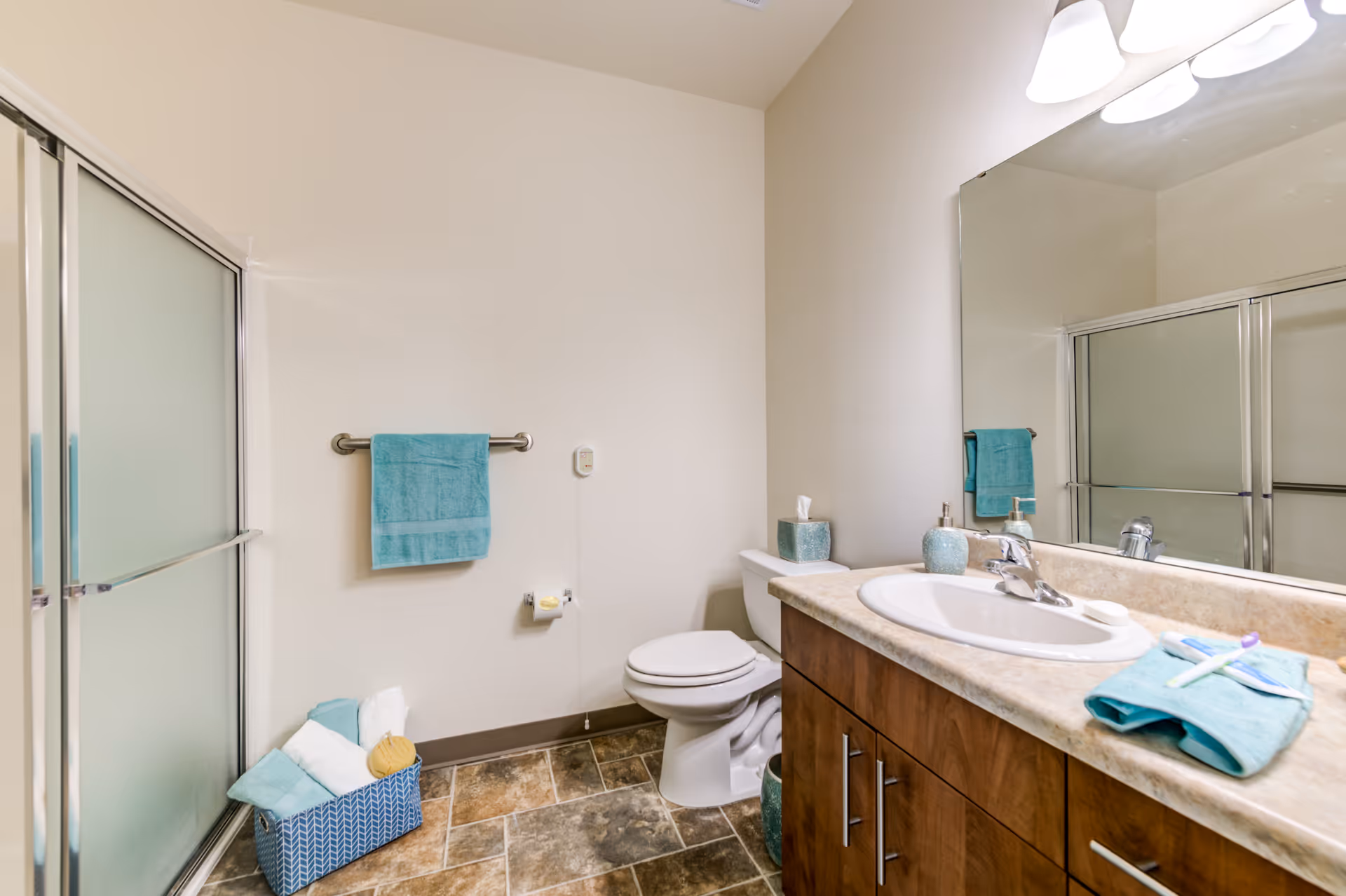 Bright bathroom featuring a shower with frosted sliding doors, a toilet, wooden vanity with sink and mirror, and teal towels and accessories.