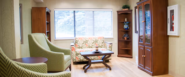 A cozy living room area with two green patterned armchairs, a floral-patterned loveseat, a wooden coffee table, wooden bookshelves, and a large window with blinds letting in natural light.