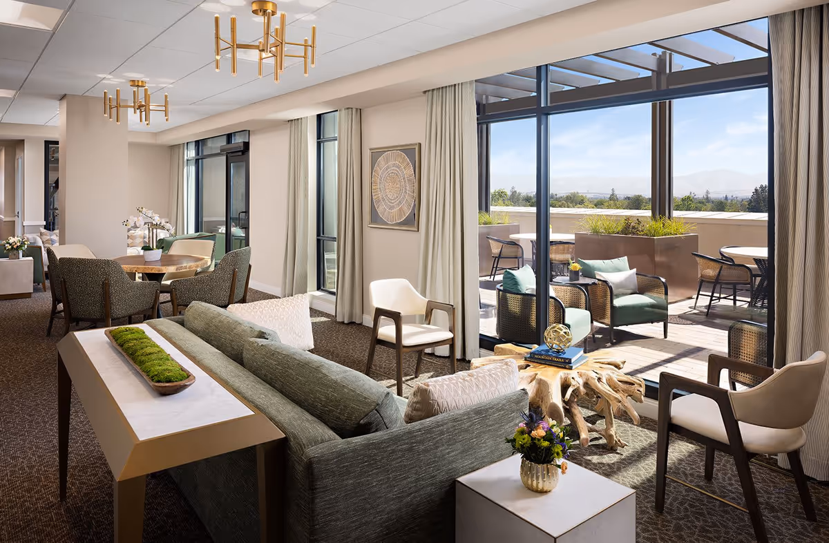 Bright, well-furnished senior living lounge with sofas, chairs, a dining area and sliding glass doors leading to a sunny outdoor terrace with seating.