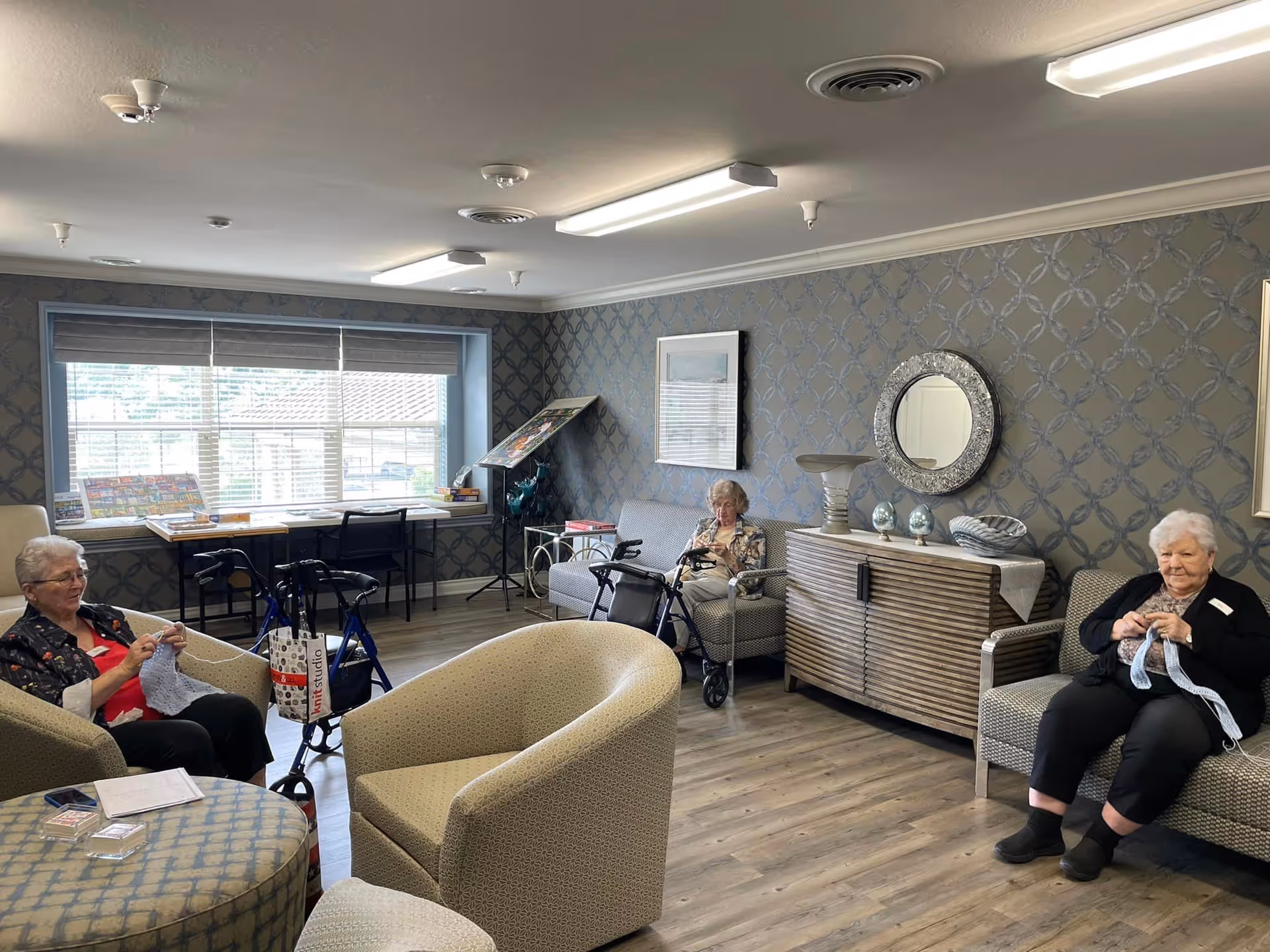 A cozy common room in a senior living facility with three elderly women sitting on armchairs and sofas, engaged in knitting. The room has patterned wallpaper, a large window with blinds, a wooden floor, and a sideboard with decorative items and a round mirror above it.