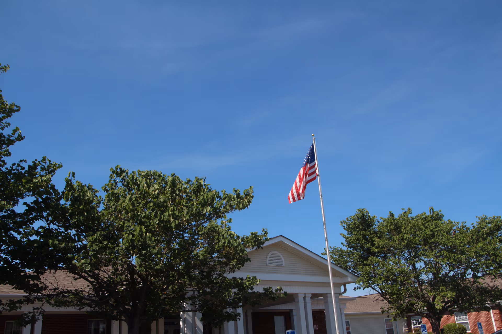 Exterior view of Wellington Manor Assisted Living building with a clear blue sky, an American flag on a flagpole, and green trees in front of the building.