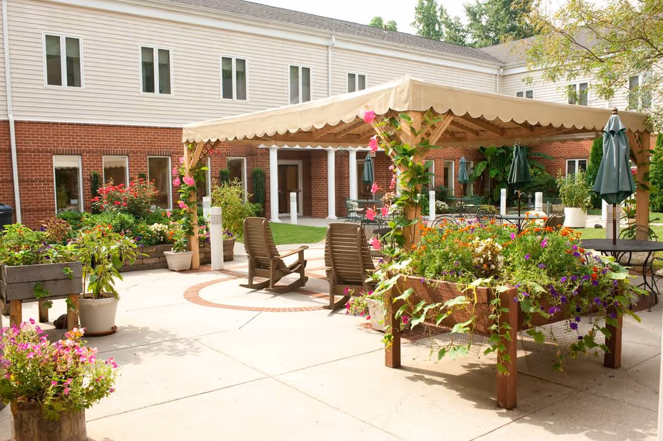 Sunny outdoor courtyard with a wooden pergola, potted flowers, lounge chairs, and a brick-and-siding building in the background.