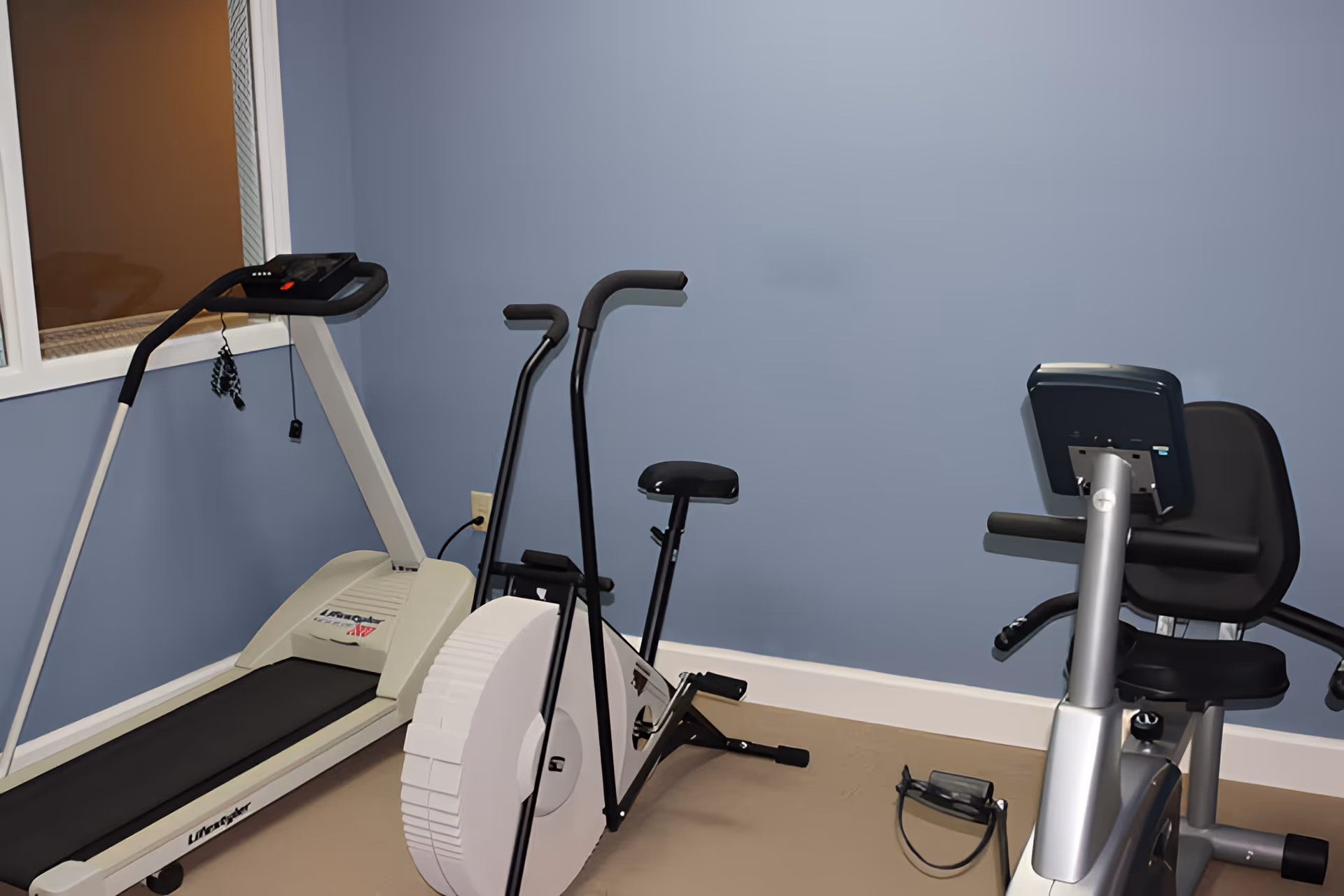A small exercise room with a treadmill, a stationary exercise bike, and a recumbent exercise bike against light blue walls and beige flooring.