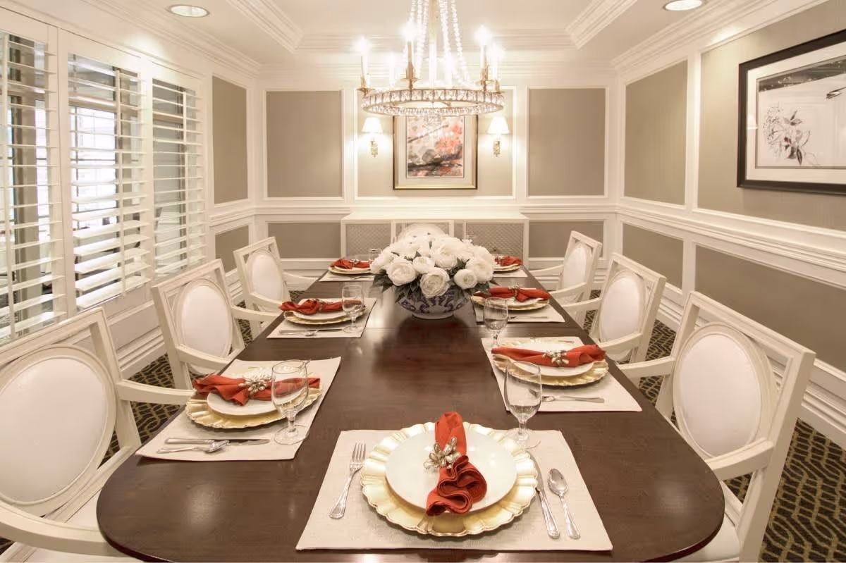 Elegant dining room with a long dark wooden table set for six people. Each place setting includes a white plate on a gold charger, a red cloth napkin with a decorative napkin ring, silverware, and a water glass. A large floral centerpiece with white flowers is in the middle of the table. The room has beige walls with white wainscoting, two framed artworks, a chandelier, and wall sconces providing warm lighting. White plantation shutters cover the windows on the left side.