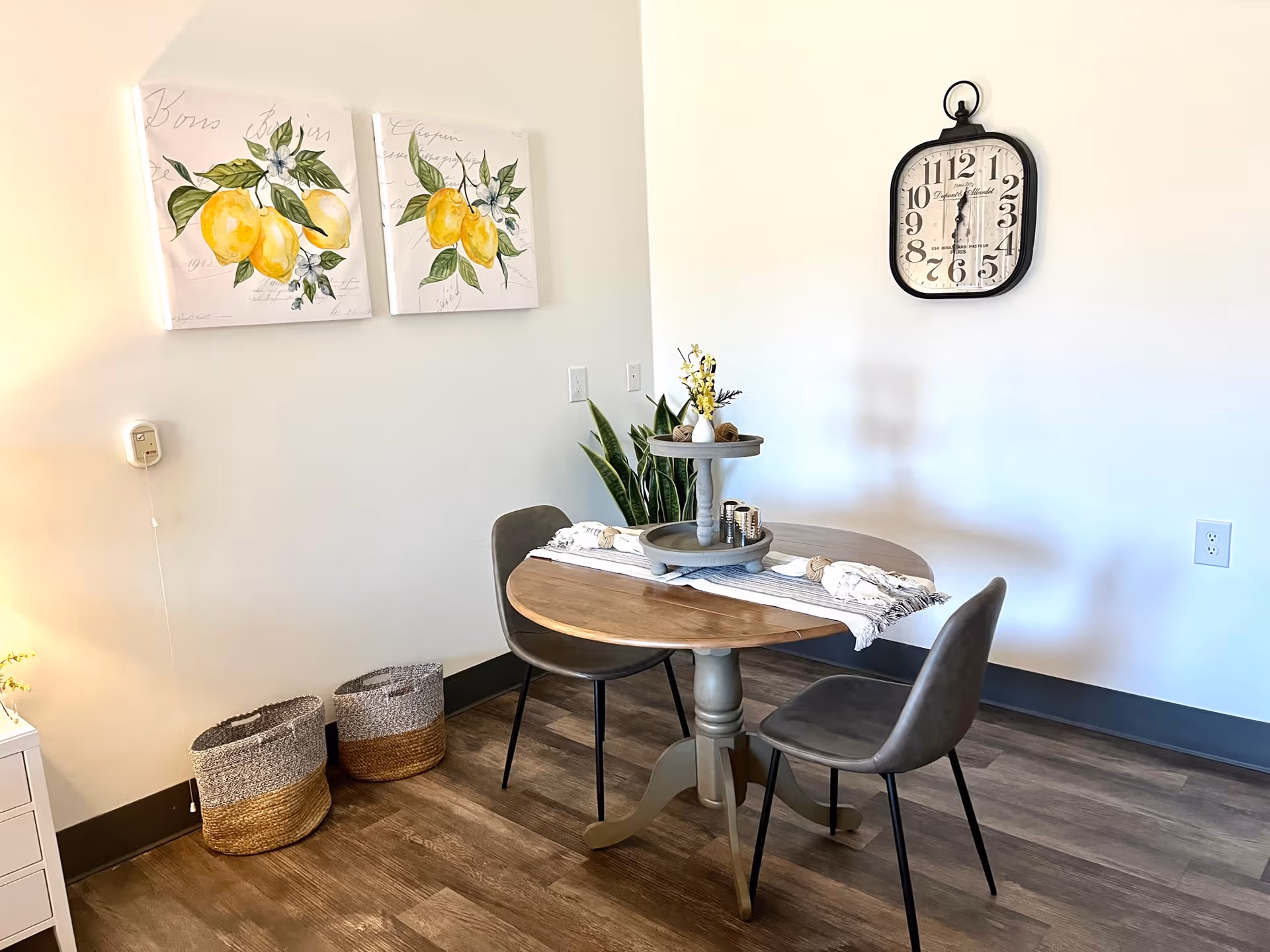Small dining nook with a round wooden table, two gray chairs, a wall clock, and lemon artwork on a light-colored wall.