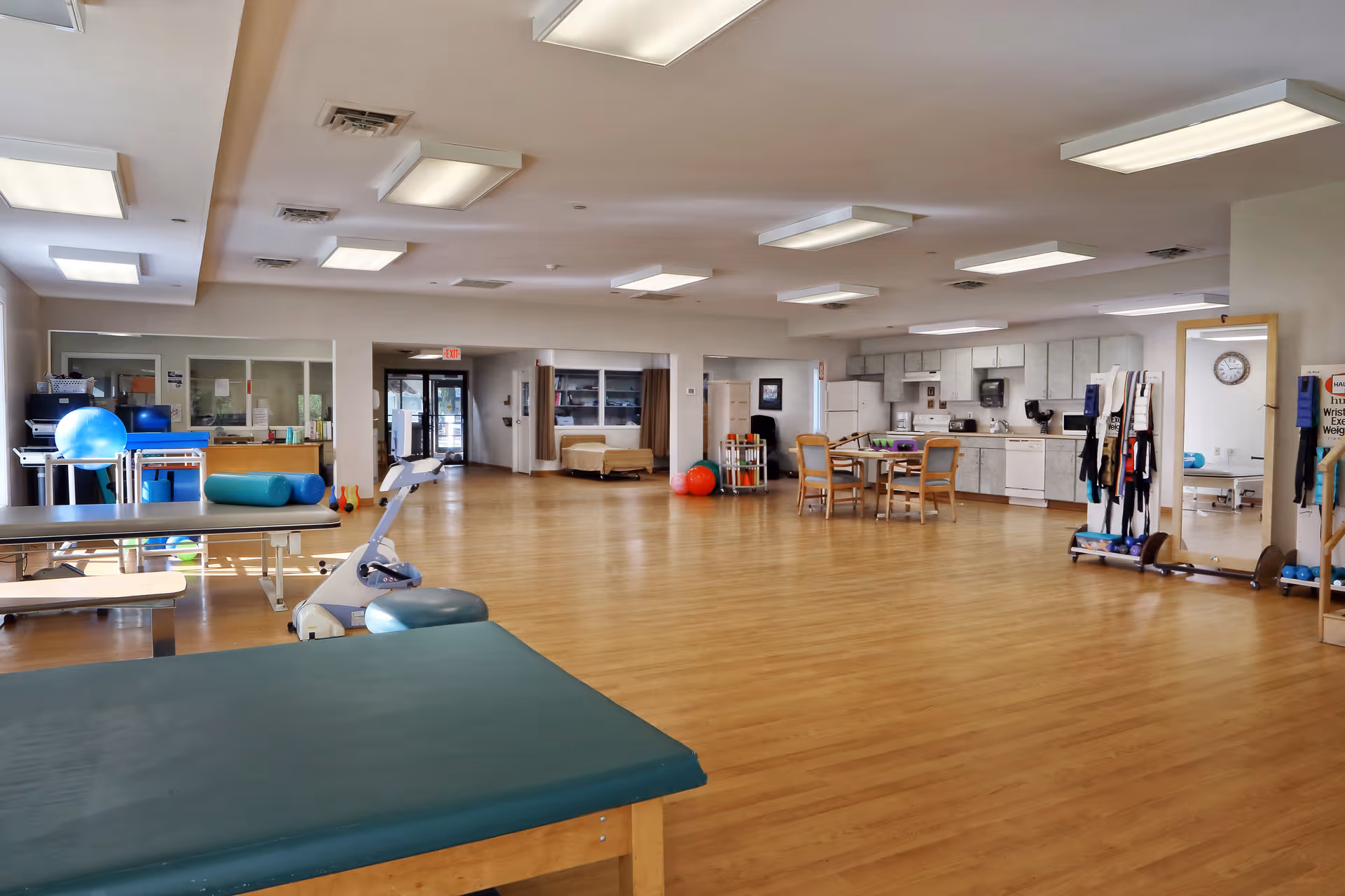 Large open therapy/activity room with exercise tables, therapy equipment, seating and a kitchenette in the background.