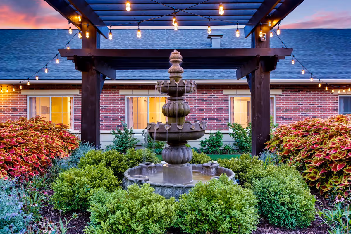 Outdoor garden area at dusk with a multi-tiered stone fountain surrounded by green bushes and colorful plants. Overhead string lights hang from a wooden pergola structure, and a brick building with lit windows is visible in the background.
