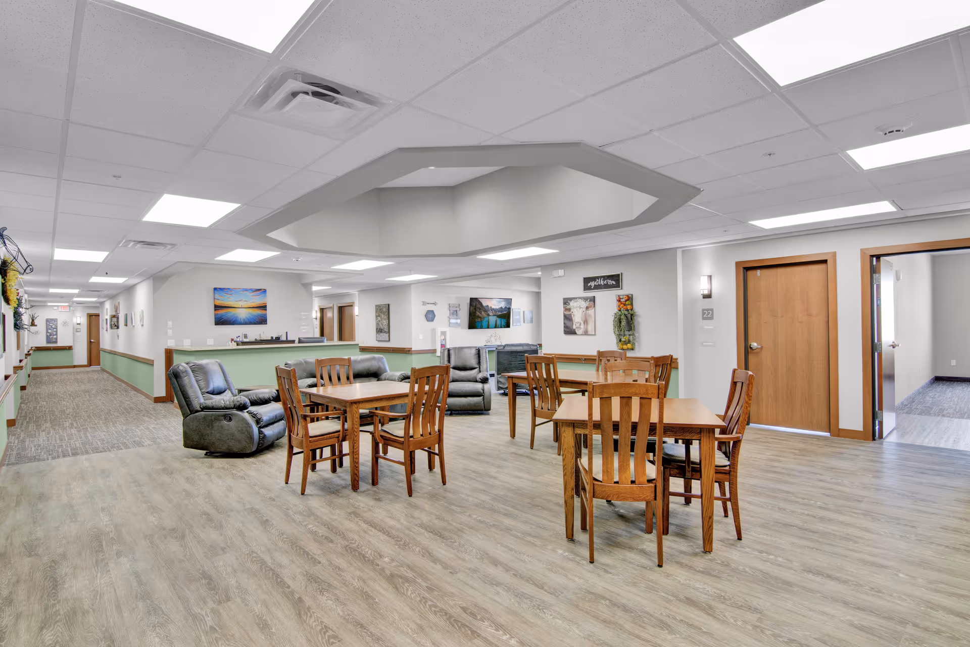 A spacious common area in a senior living facility featuring wooden tables and chairs, two gray recliner chairs, and a green reception desk. The room has light-colored walls with artwork and decorations, a ceiling with recessed lighting, and a hallway leading to other rooms.