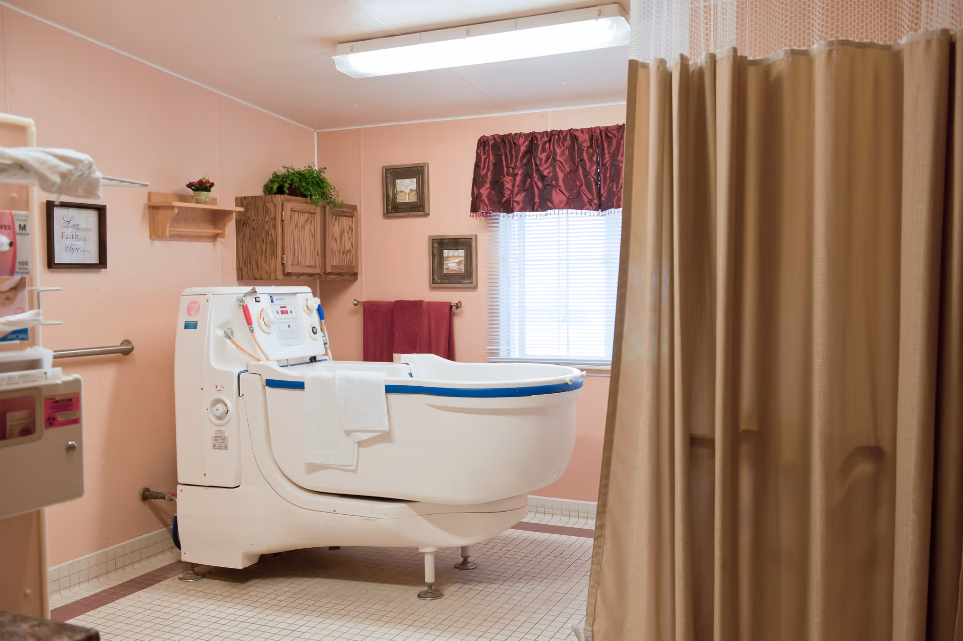A bathroom with a large white walk-in bathtub equipped with controls and a towel hanging over the side. The walls are painted light pink, and there are wooden cabinets and framed pictures on the wall. A window with blinds and a burgundy valance is visible, along with a beige privacy curtain partially drawn on the right side.