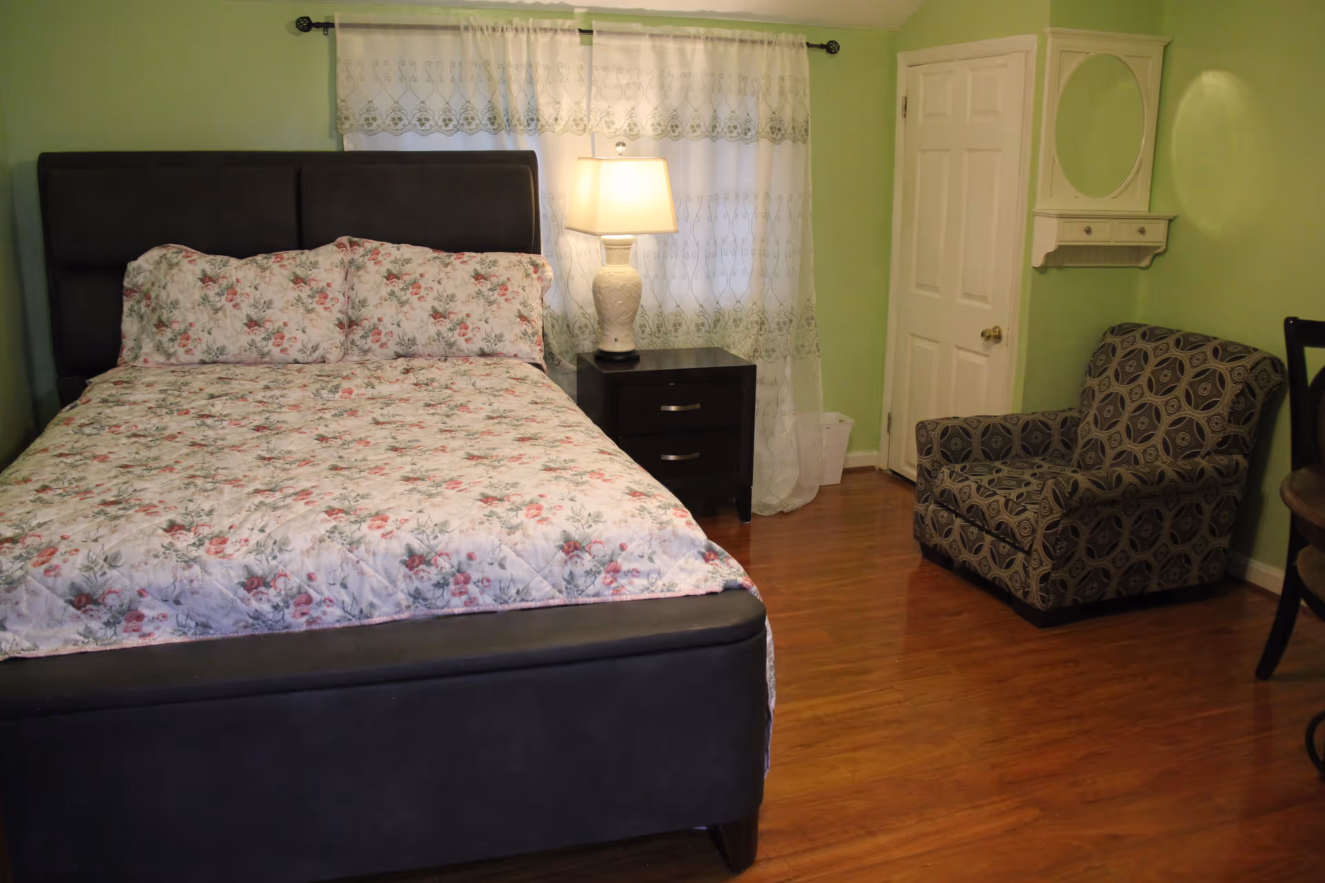 A bedroom with a dark upholstered bed frame and floral bedding, a nightstand with a white lamp, a patterned armchair, a white door, a wall-mounted mirror with a small shelf, and light green walls with wood flooring.