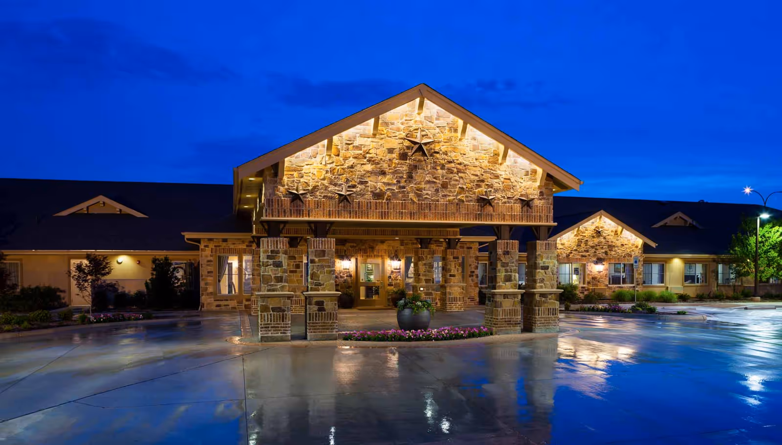 Exterior front view of Meadowbrook Memory Care Community building at dusk, featuring a stone facade with illuminated star decorations, a covered entrance supported by stone pillars, and a wet driveway reflecting the building lights.
