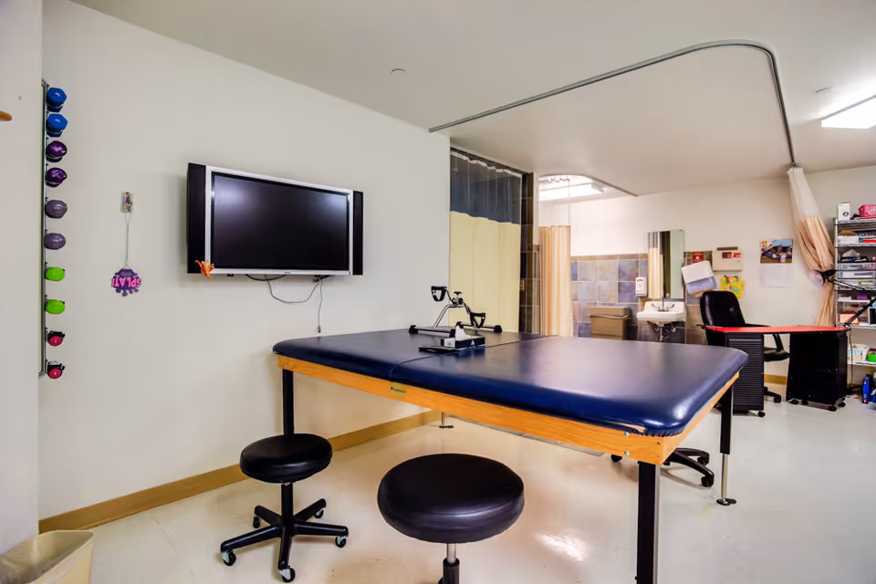 A therapy room with a padded treatment table, two black stools on wheels, a wall-mounted TV, and exercise equipment on the wall. In the background, there is a sink, a chair, shelves with supplies, and a privacy curtain on a ceiling track.