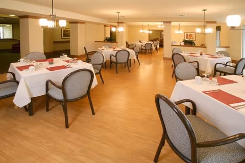 A spacious dining room with multiple tables covered in white tablecloths and set with red placemats, cups, and utensils. The room has wooden flooring, cushioned chairs with armrests, and warm lighting from ceiling fixtures. There are plants and framed artwork on the walls, creating a welcoming atmosphere.