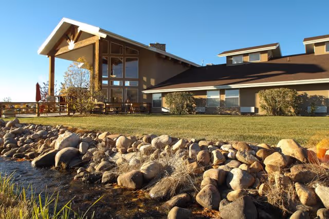 Exterior view of Elk Meadows Assisted Living & Memory Care building with a sloped roof, large windows, and a covered porch area. In the foreground, there is a landscaped area with rocks, grass, and a small water feature under a clear blue sky.
