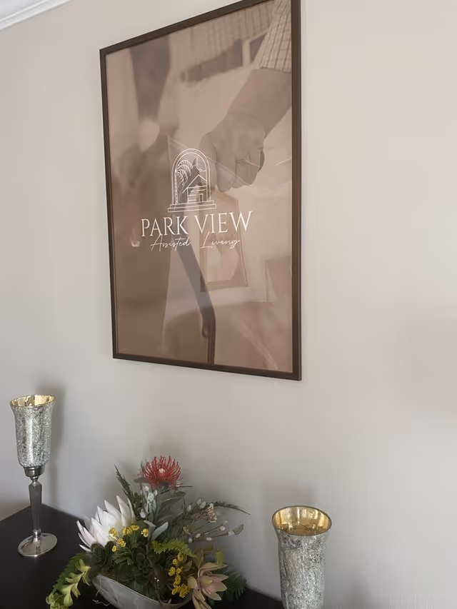 A framed poster on a wall with the text 'PARK VIEW Assisted Living' featuring an illustration of a house and palm trees. Below the poster is a dark surface with a floral arrangement and two decorative silver candle holders.