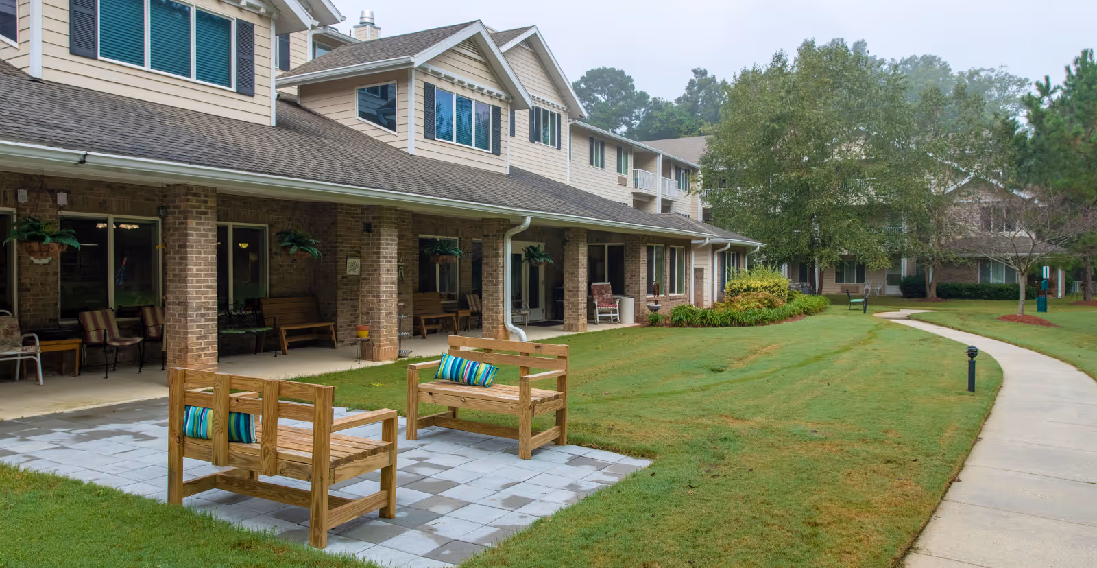 Outdoor patio area at Jordan Oaks Gracious Retirement Living featuring wooden benches with colorful cushions on a tiled section, a covered porch with brick pillars and hanging plants, surrounded by green grass and trees with a curved concrete walkway.