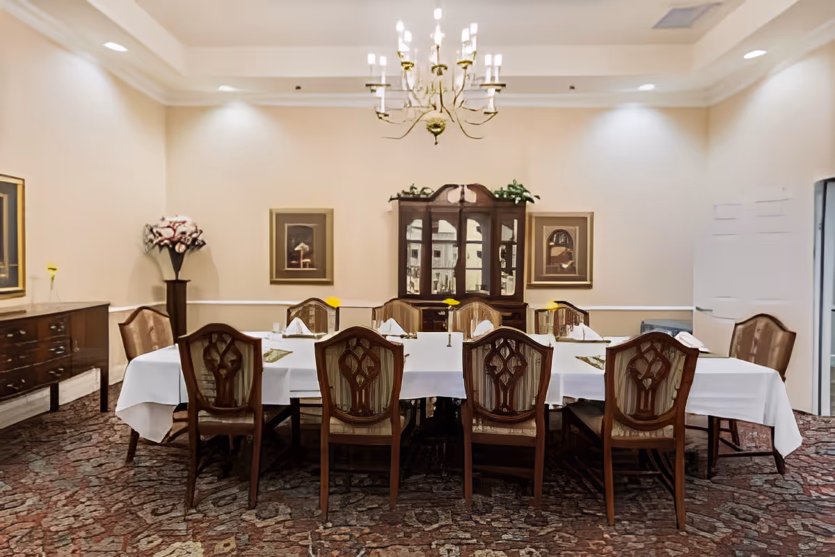 A formal dining room with a long rectangular table covered with a white tablecloth, surrounded by wooden chairs with patterned upholstery. The table is set with folded napkins and small yellow flowers in vases. A wooden china cabinet with glass doors is against the back wall, flanked by two framed paintings. A chandelier hangs from the ceiling, and the room has beige walls and a patterned carpet.