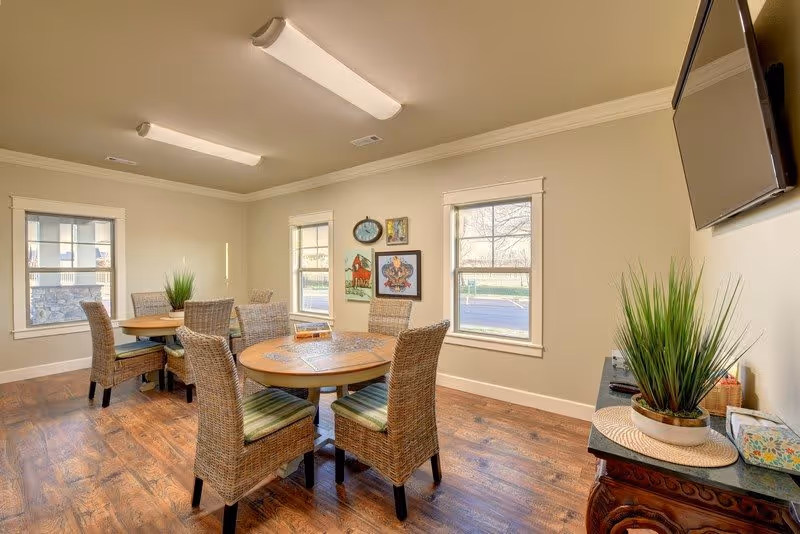 A bright room with wooden flooring featuring two round tables surrounded by wicker chairs with green cushions. The walls are light-colored with three windows letting in natural light. There are framed pictures and a clock on one wall, a flat-screen TV mounted on another wall, and a small table with a potted plant and tissue box underneath the TV.