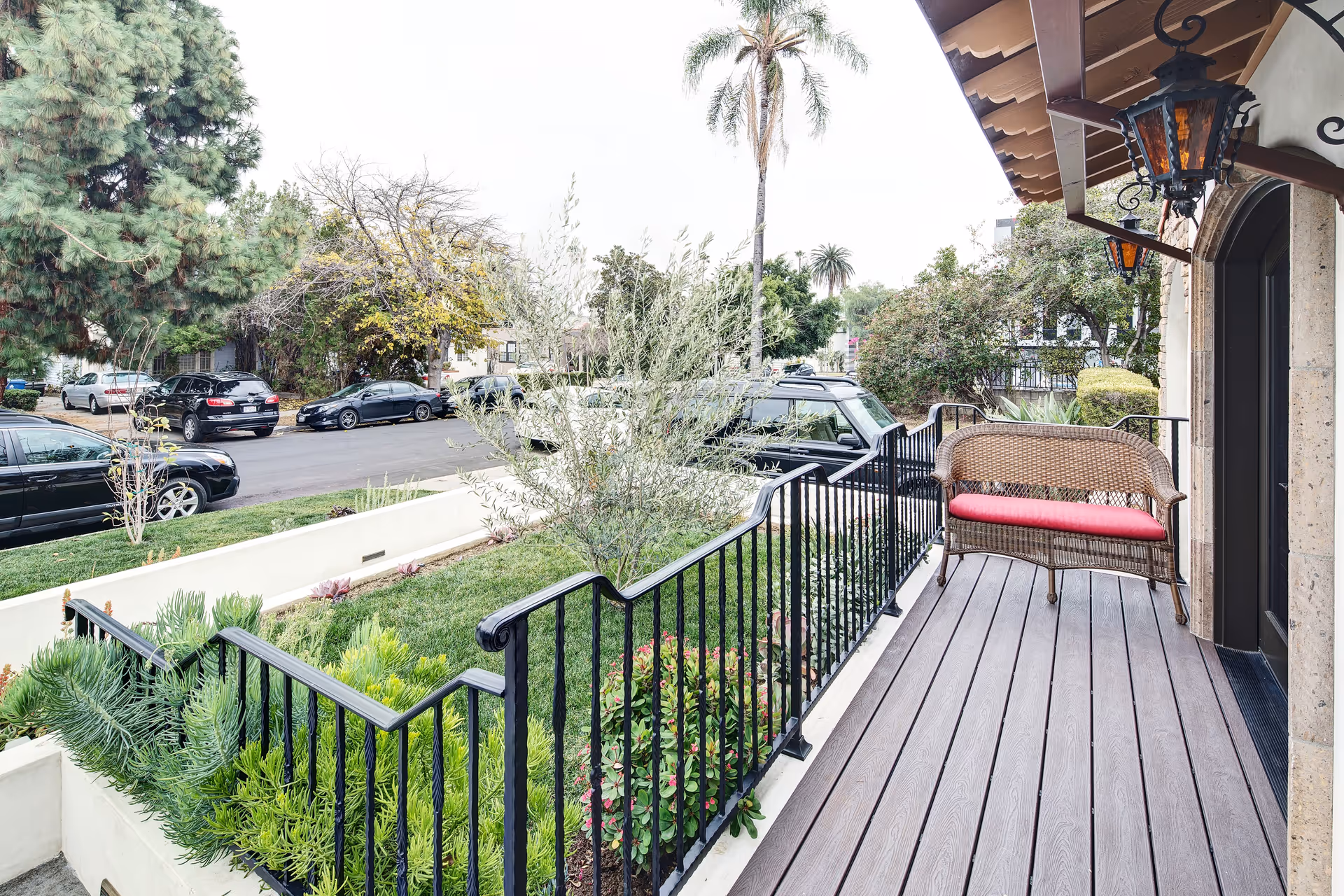 A front porch area with a wooden floor, black metal railing, and a wicker bench with a red cushion. There are two decorative lantern-style lights hanging from the porch ceiling. The porch overlooks a small garden with various green plants and shrubs. In the background, there is a street with parked cars and trees, including a tall palm tree.